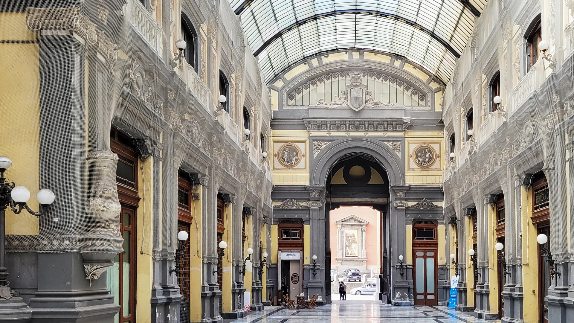 Uvnitř Gallerie Principe di Napoli: dlouhá skleněná a železná klenutá pasáž s malovanými žlutými a šedými fasádami a mramorovou podlahou